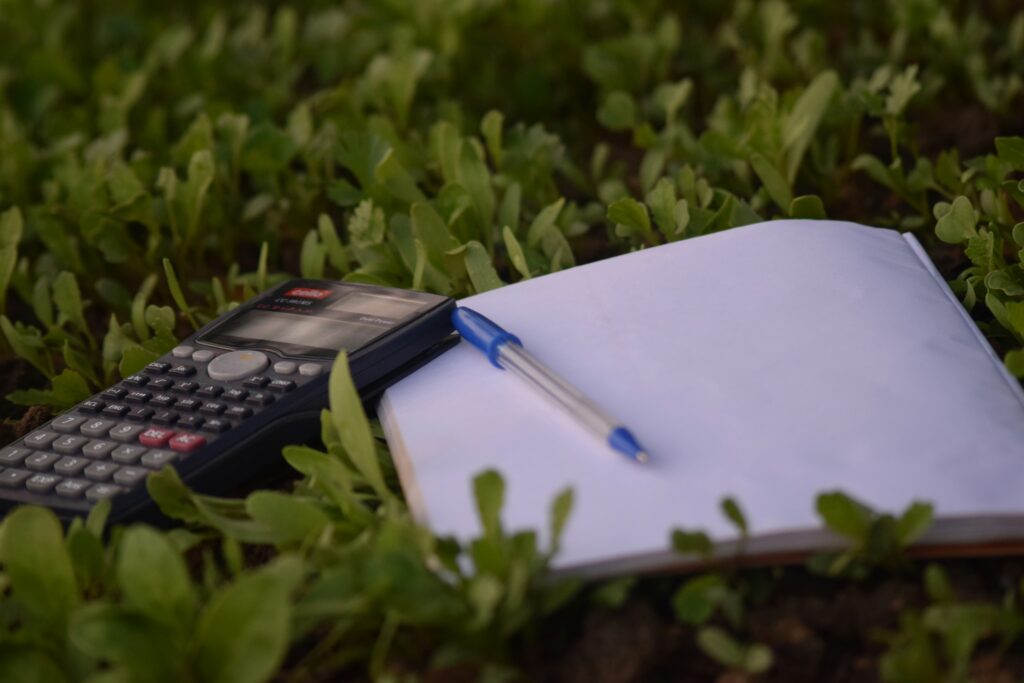 A calculator, blue pen, and blank notebook resting on green grass, symbolizing budgeting, planning, and financial reflection in a peaceful outdoor setting.
