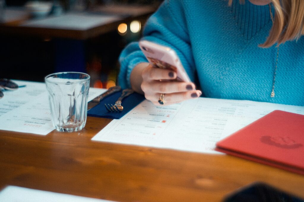 Person in a blue sweater sitting at a table, checking a smartphone while looking at a restaurant menu — symbolizing everyday spending decisions and mindful budgeting.
