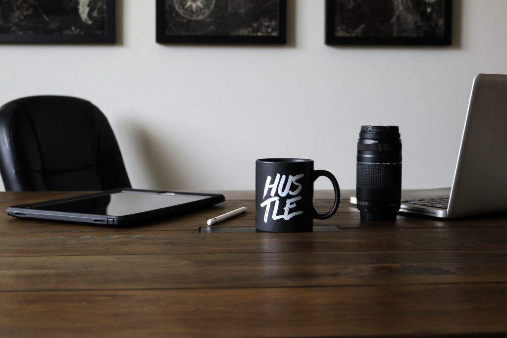 Minimalist wooden desk setup with a black coffee mug labeled “Hustle,” next to a laptop, camera lens, and tablet — symbolizing focus, productivity, and side hustling.