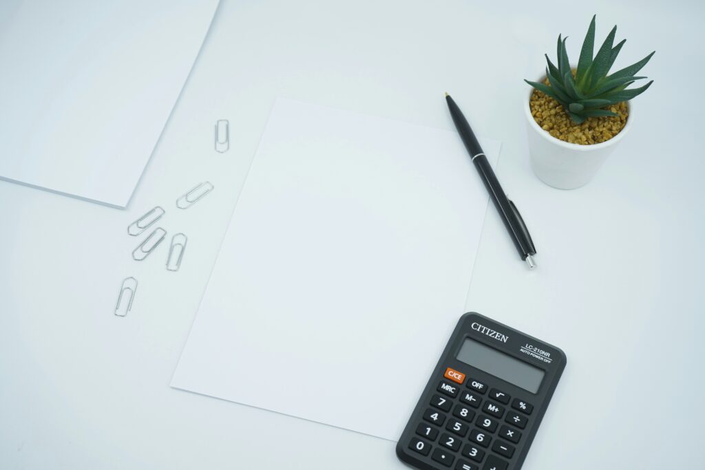 Minimalist desk setup with blank paper, a black pen, scattered paper clips, a small potted succulent, and a calculator — representing budgeting or financial planning.