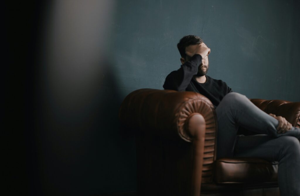 A man sitting on a brown leather couch with his hand over his face, looking stressed or overwhelmed.