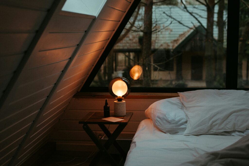A warm, softly lit bedroom with a glowing bedside lamp, a wooden nightstand, and a cozy unmade bed beneath a slanted ceiling and large window at dusk